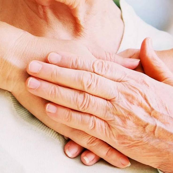 Hände aufs Herz - stehende junge Person legt von hinten einem sitzenden älteren Menschen die Hände herzseitig auf der Brust übereinander, und der ältere Mensch legt seine linke Hand auf die jungen Hände