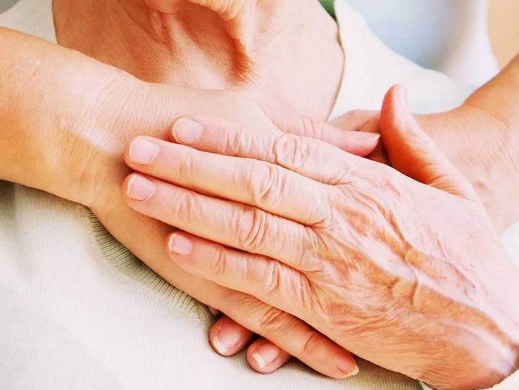 Hände aufs Herz - stehende junge Person legt von hinten einem sitzenden älteren Menschen die Hände herzseitig auf der Brust übereinander, und der ältere Mensch legt seine linke Hand auf die jungen Hände