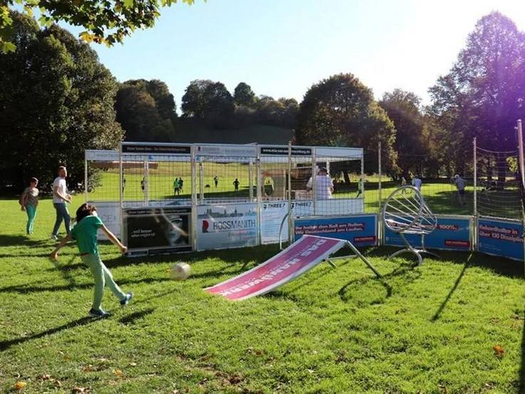 Kinder spielen im Soccer Cage und an der Fußballrampe mit Fangkorb des auf sonniger Wiese im Freien aufgebauten Funparks