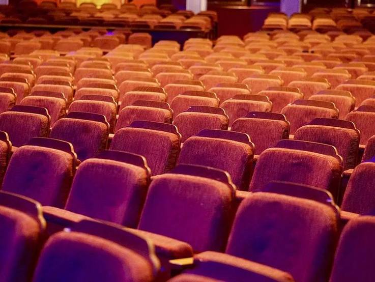 Bildausschnitt rot gepolsterter Theaterstuhlreihen in etwas abgedunkeltem, künstlichem Licht