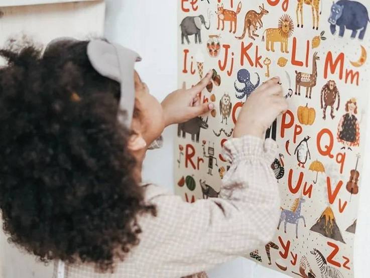 Ein Mädchen betrachtet ein Buchstabenplakat mit Tieren und zeigt auf ausgewählte Stellen des Plakats