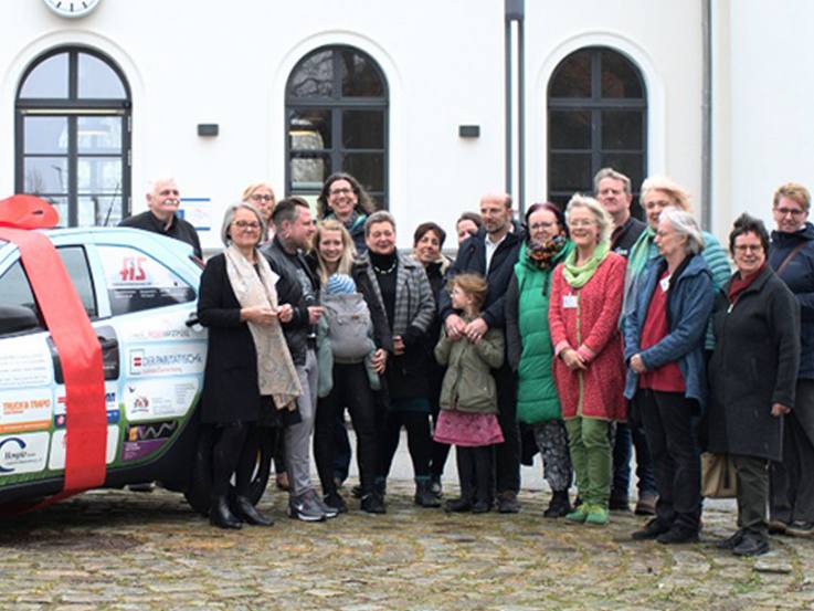 Folierter gesponsorter Toyota Aygo X mit roter Schleife darum links von den Sponsoren und Mitwirkenden des Hospizvereins Lüchow-Dannenberg