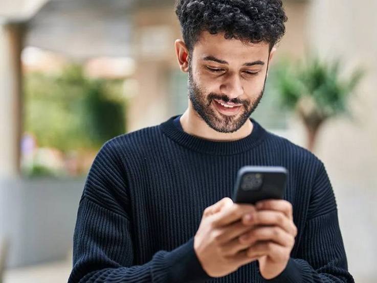 Junger Mann, der auf seinen Mobildtelefonbildschirm schaut während er das Gerät in beiden Händen hält