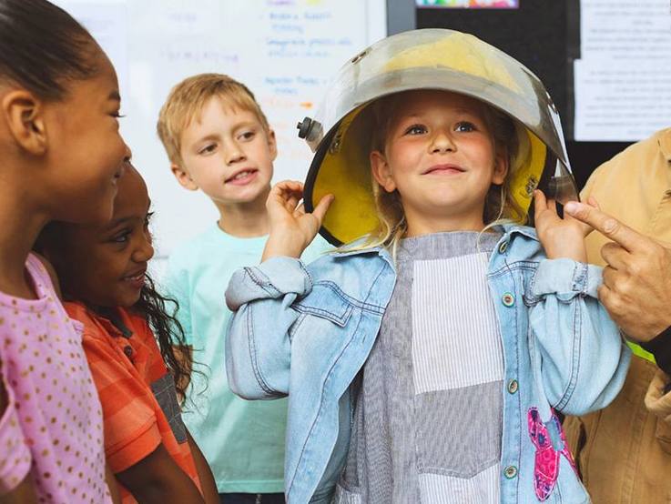 Kinderschar, deren Aufmerksamkeit auf ein Mädchen in der Mitte gerichtet ist, die gerade einen viel zu großen Feuerwehrschutzhelm anprobiert