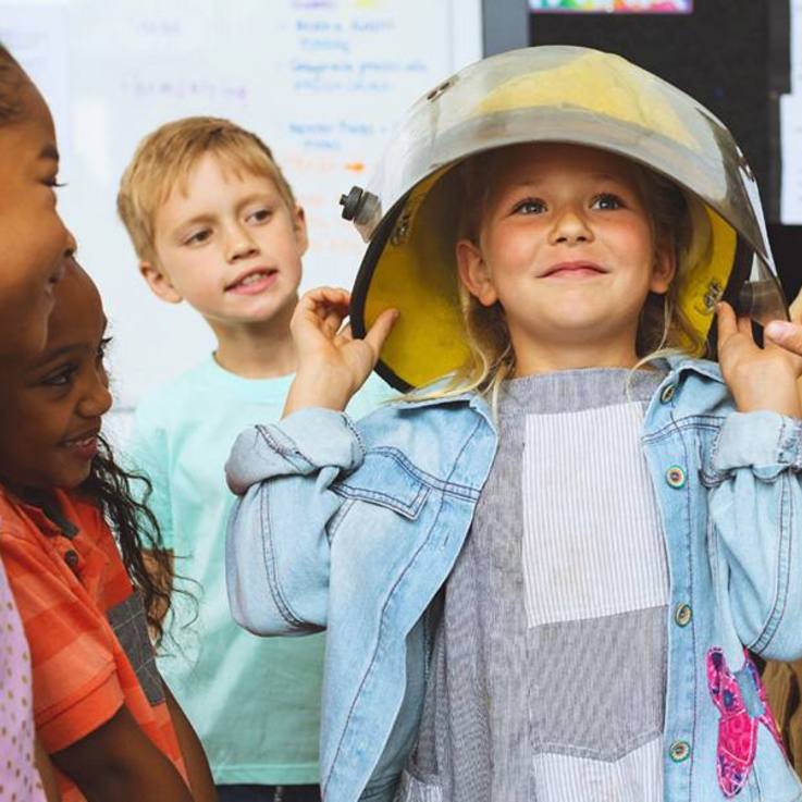 Kinderschar, deren Aufmerksamkeit auf ein Mädchen in der Mitte gerichtet ist, die gerade einen viel zu großen Feuerwehrschutzhelm anprobiert