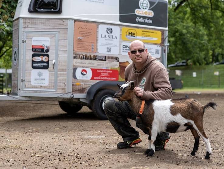 Manuel Ferkau, 1. Vorsitzender des Tierschutzvereins Hattersheim, neben dem folierten gesponsorten Tiertransportanhänger kniend - eine braun-weiße Ziege an seiner Seite