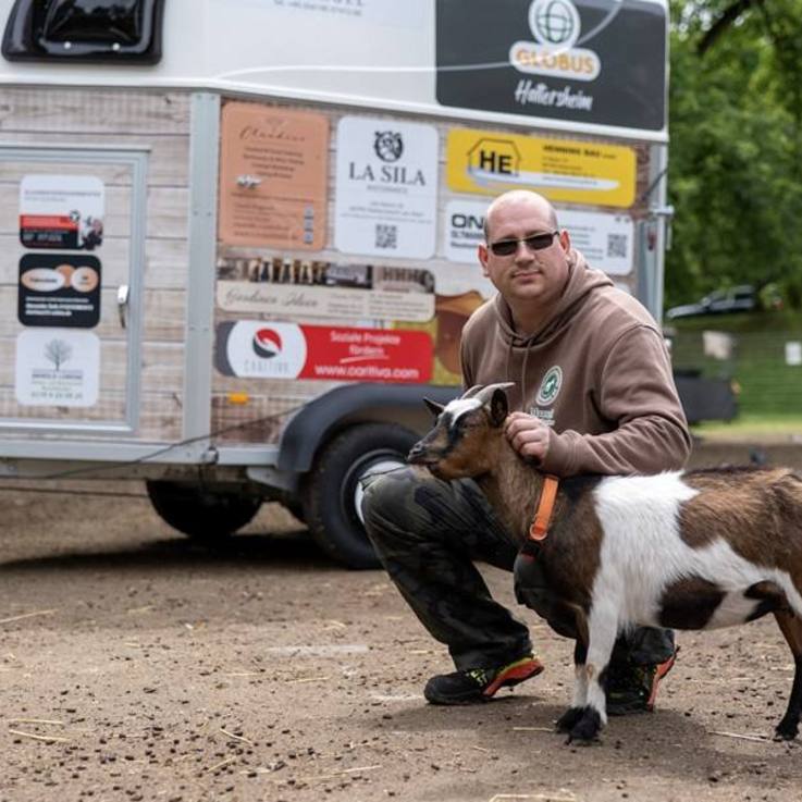 Manuel Ferkau, 1. Vorsitzender des Tierschutzvereins Hattersheim, neben dem folierten gesponsorten Tiertransportanhänger kniend - eine braun-weiße Ziege an seiner Seite